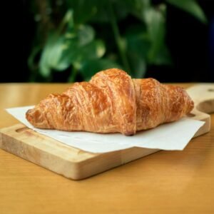 Golden-brown croissant placed on a wooden board inside a café, perfect for breakfast or snack time.